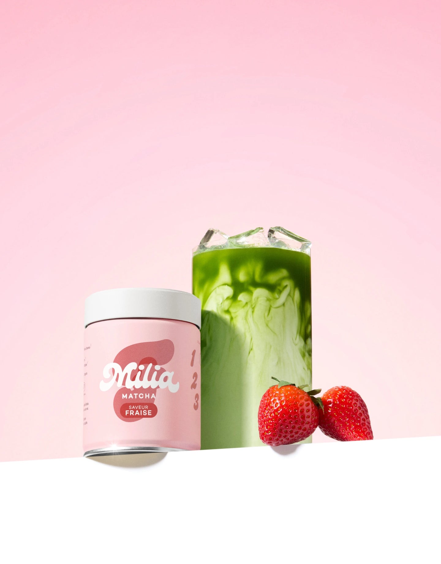 Milia Matcha Matcha-ceremoniële blikje Milia Matcha , met ijskoude matcha latte en twee aardbeien op een roze achtergrond