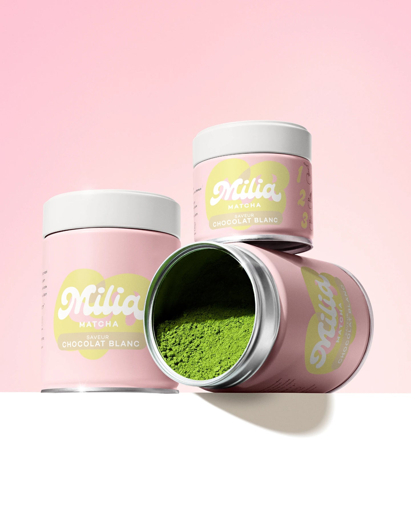 Trois canettes de matcha Milia Matcha saveur chocolat blanc dont une ouverte révélant poudre de matcha verte sur fond rose